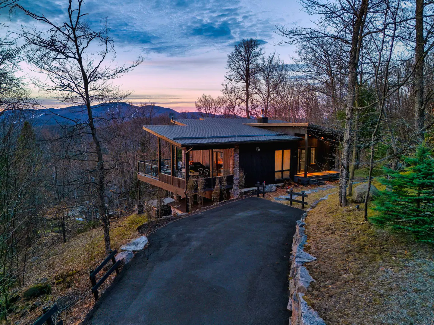 Modern dark home with a metal roof sits on a hill overlooking mountains at dusk. A paved driveway leads up to the house.