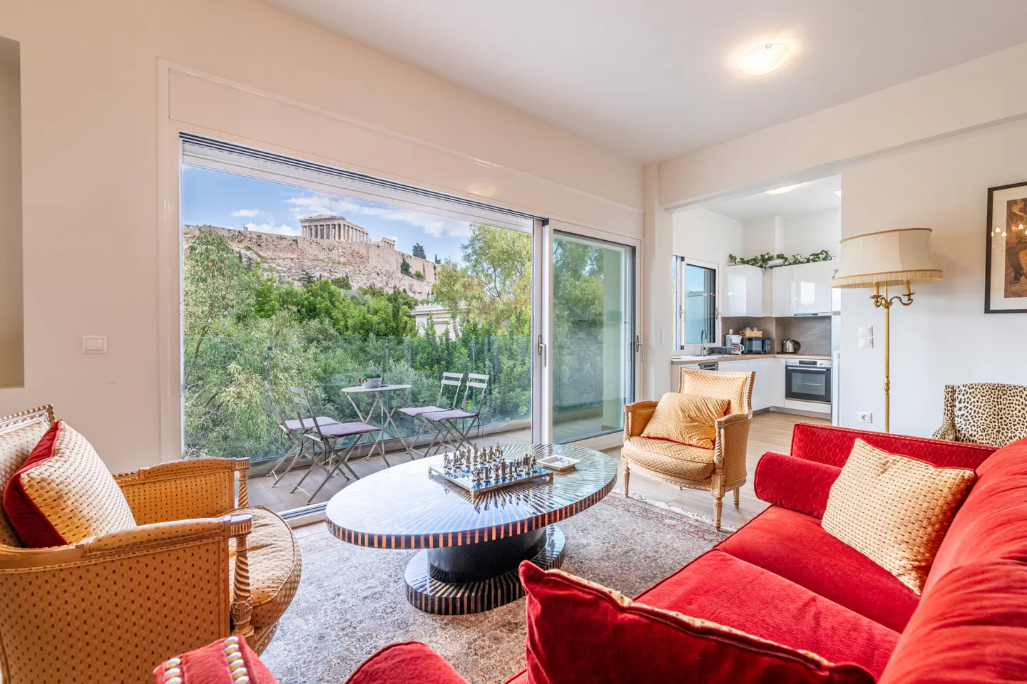 Living room with a red sofa, patterned armchairs, and a coffee table, featuring a balcony view of the Acropolis in Athens.