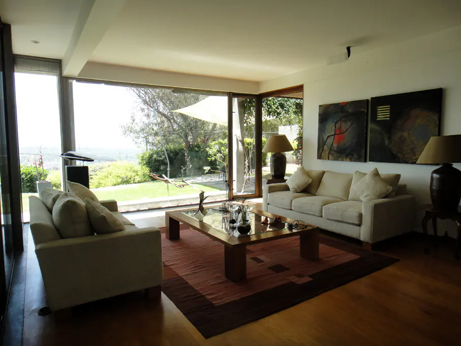 Bright living room with hardwood floors, two cream sofas, and a wood coffee table on a patterned rug. Large windows overlook a green lawn.