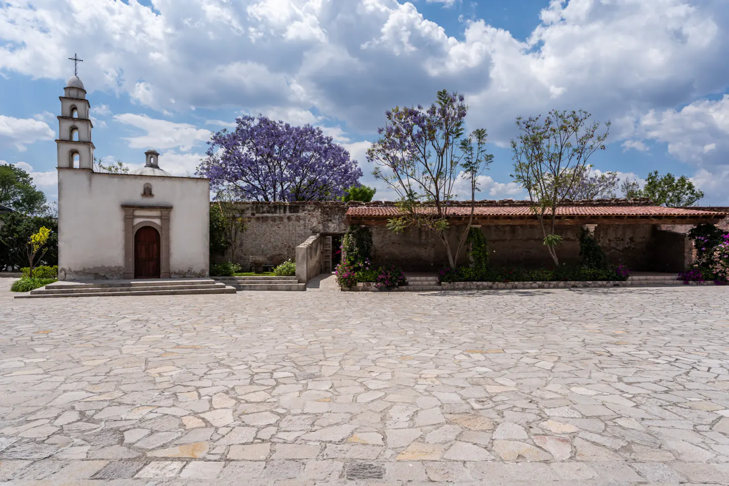 Exterior of a white chapel with a bell tower, brown door, and stone courtyard under a cloudy sky. Purple trees in the background.