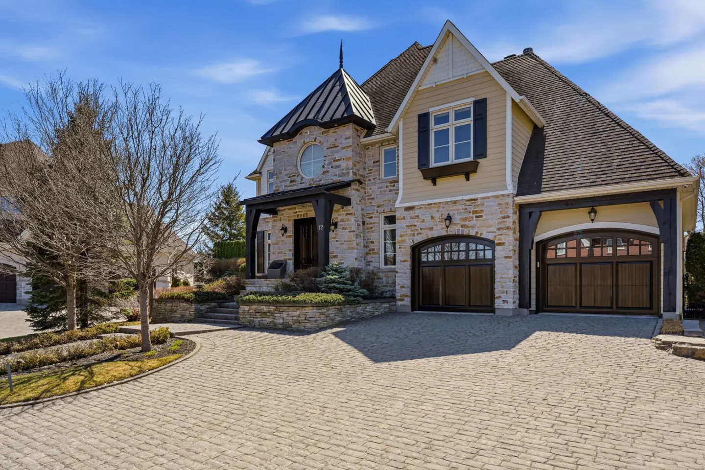 Two-story stone house with a turret, dark brown garage doors, and a cobblestone driveway under a blue sky.