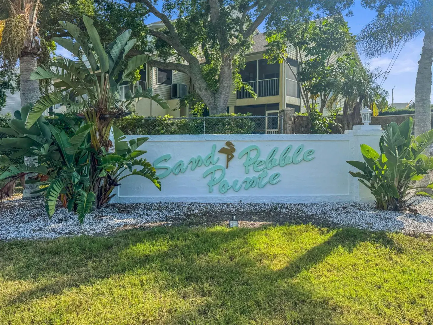 "Sand Pebble Pointe" sign in light blue on a white wall, surrounded by tropical plants and green grass. Condos are visible in the background.