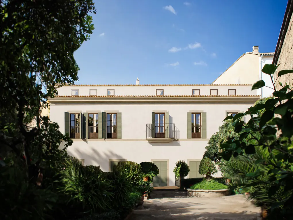 Two-story white building with green shutters, seen through lush green foliage under a blue sky. Balcony on the second floor.