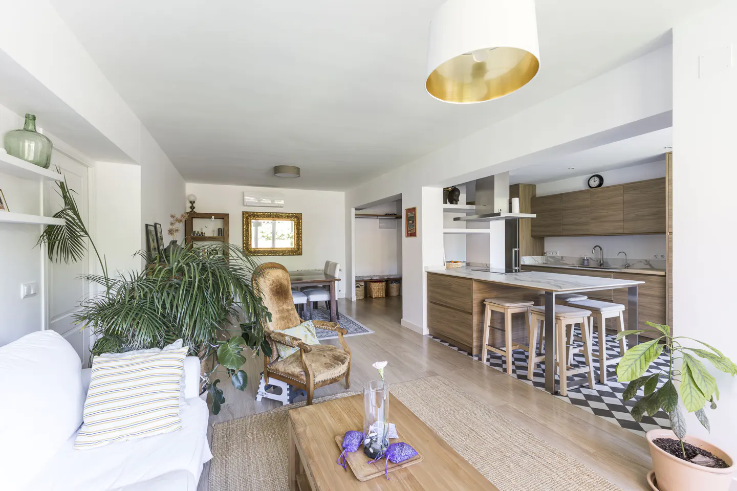 Bright, open-concept living space with a white sofa, vintage armchair, and a modern kitchen with an island and stools. Plants add a touch of nature.