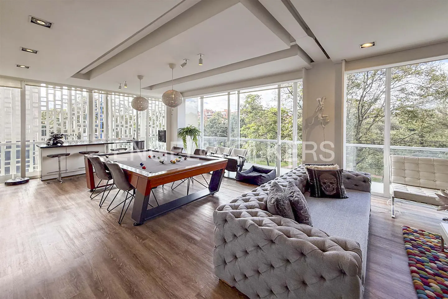 Bright, modern living room with a pool table, grey tufted sofa, and floor-to-ceiling windows overlooking greenery.