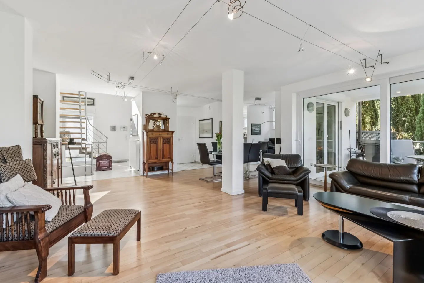 Bright, open living room with wood floors, white walls, and modern lighting. Antique wood furniture contrasts with black leather sofas. Sliding glass doors lead to a patio.