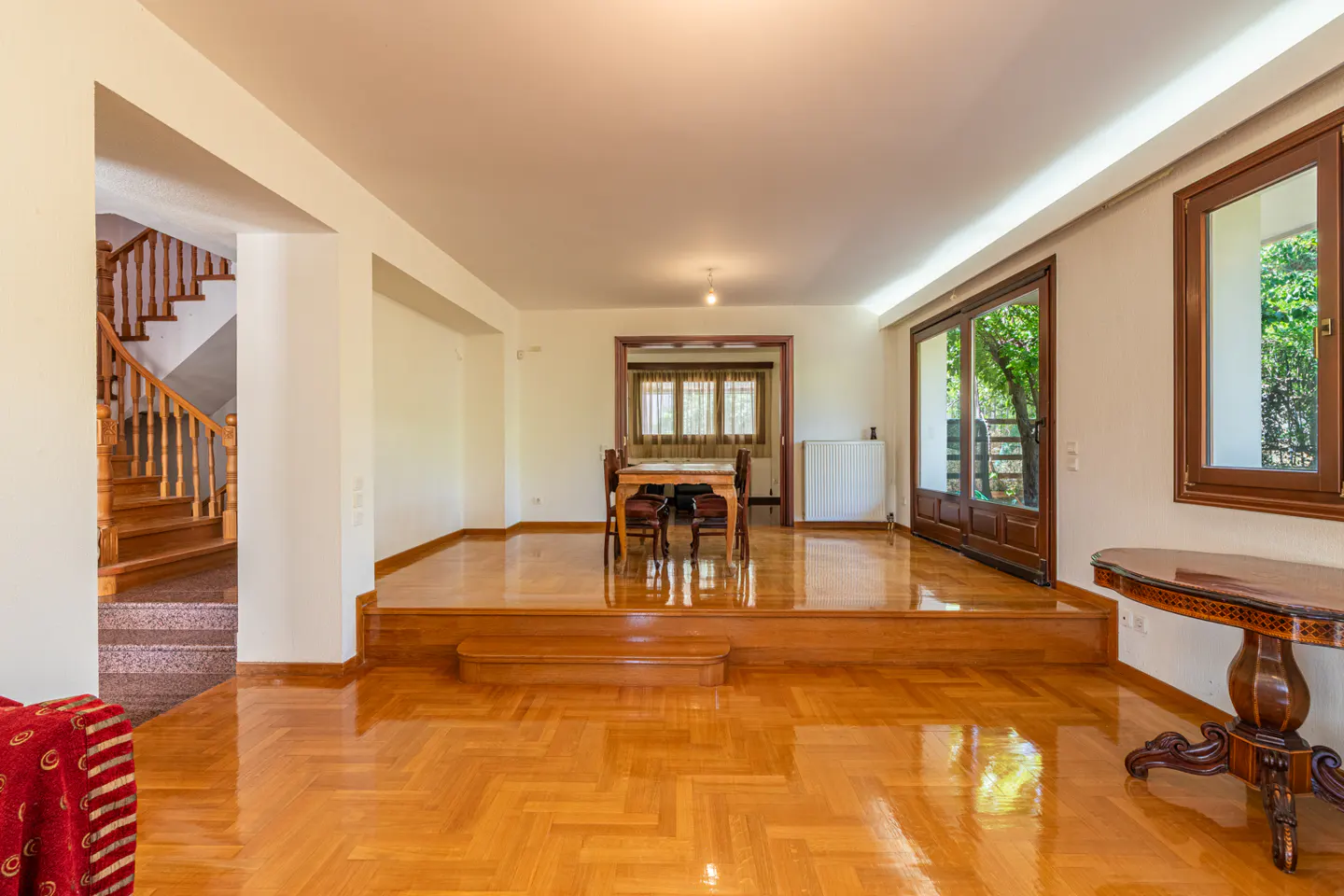 Spacious living area with polished wood floors, a raised dining room, wooden staircase, and a classic pedestal side table.