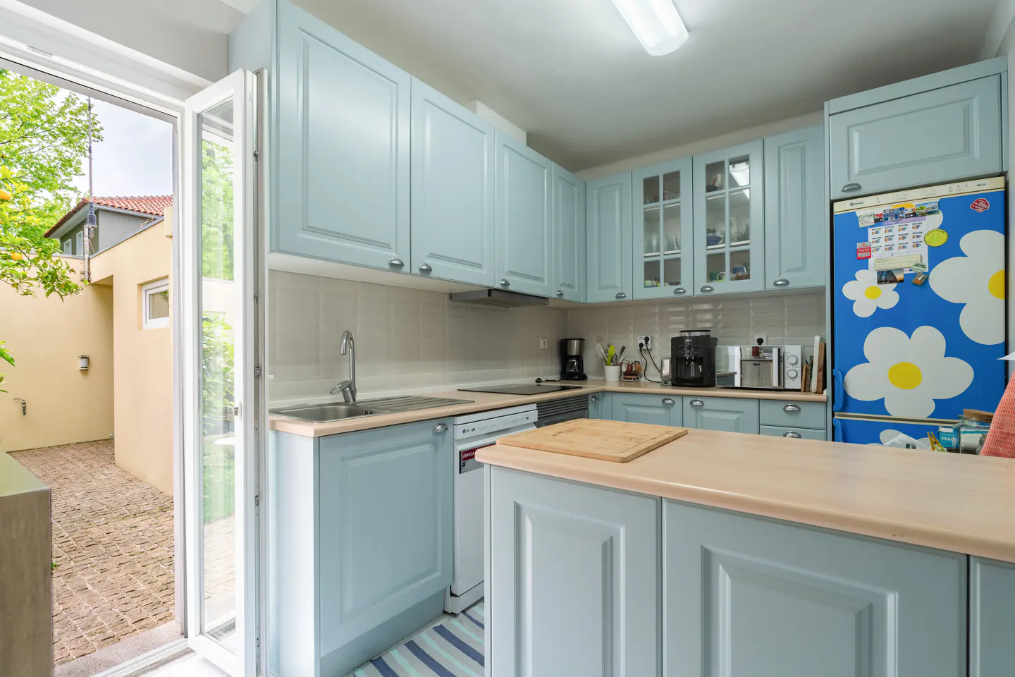 Bright kitchen with light blue cabinets, stainless steel appliances, and a butcher block countertop. An open door leads to a brick patio.