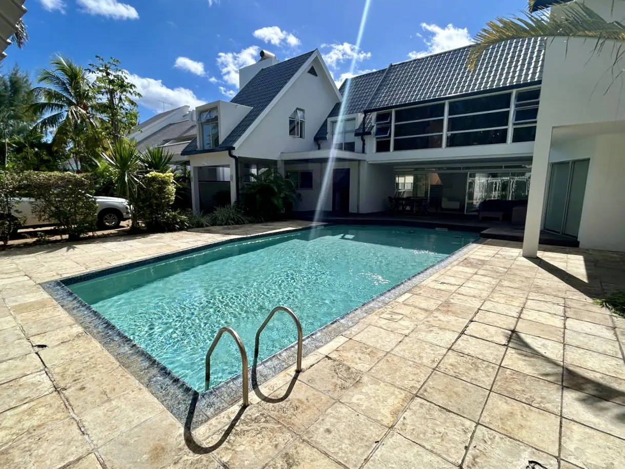 A backyard view of a white house with a pool, stone patio, and blue sky.