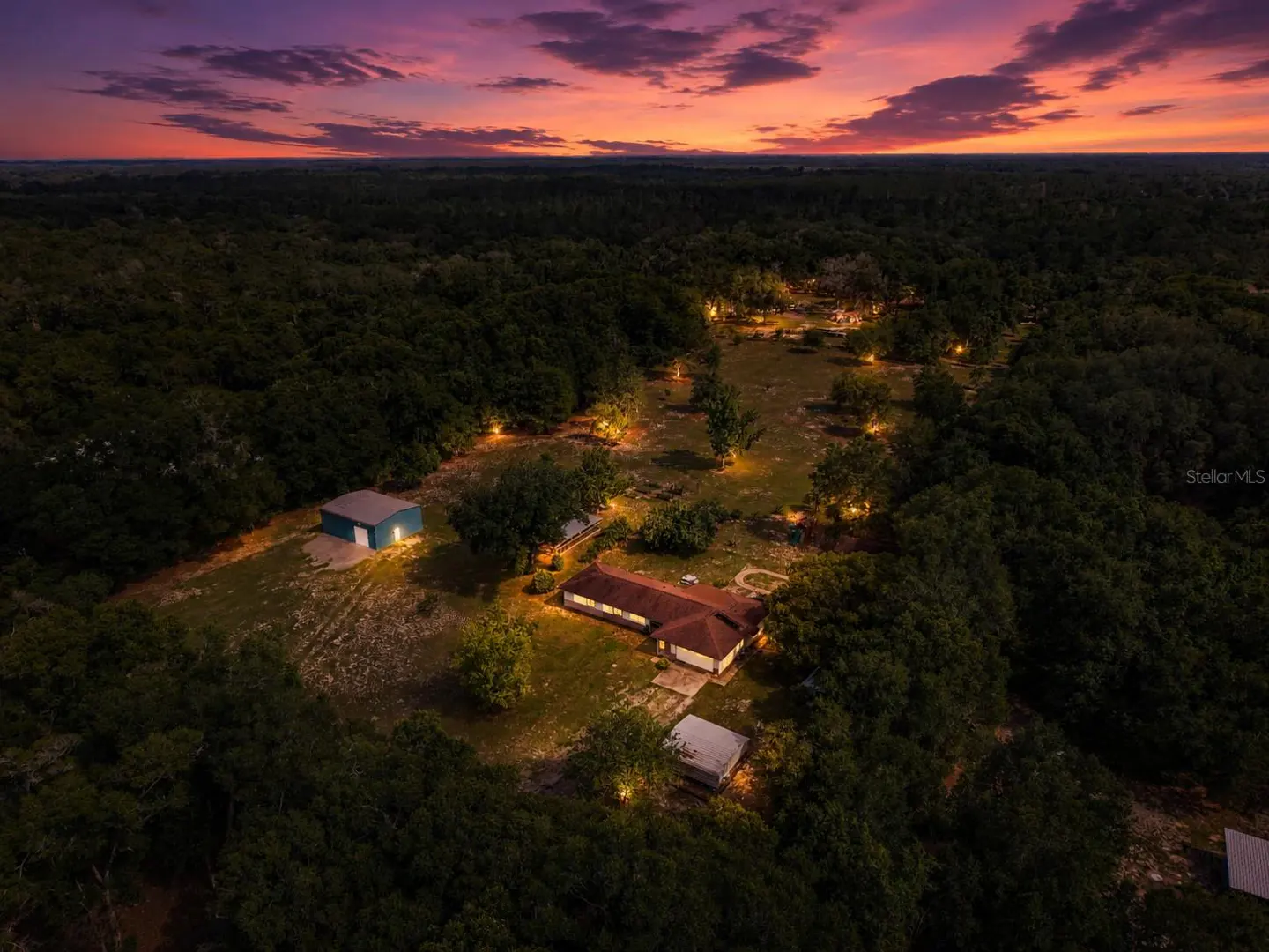 Aerial view of a rural property with a house and outbuildings illuminated at dusk under a vibrant purple and orange sky.