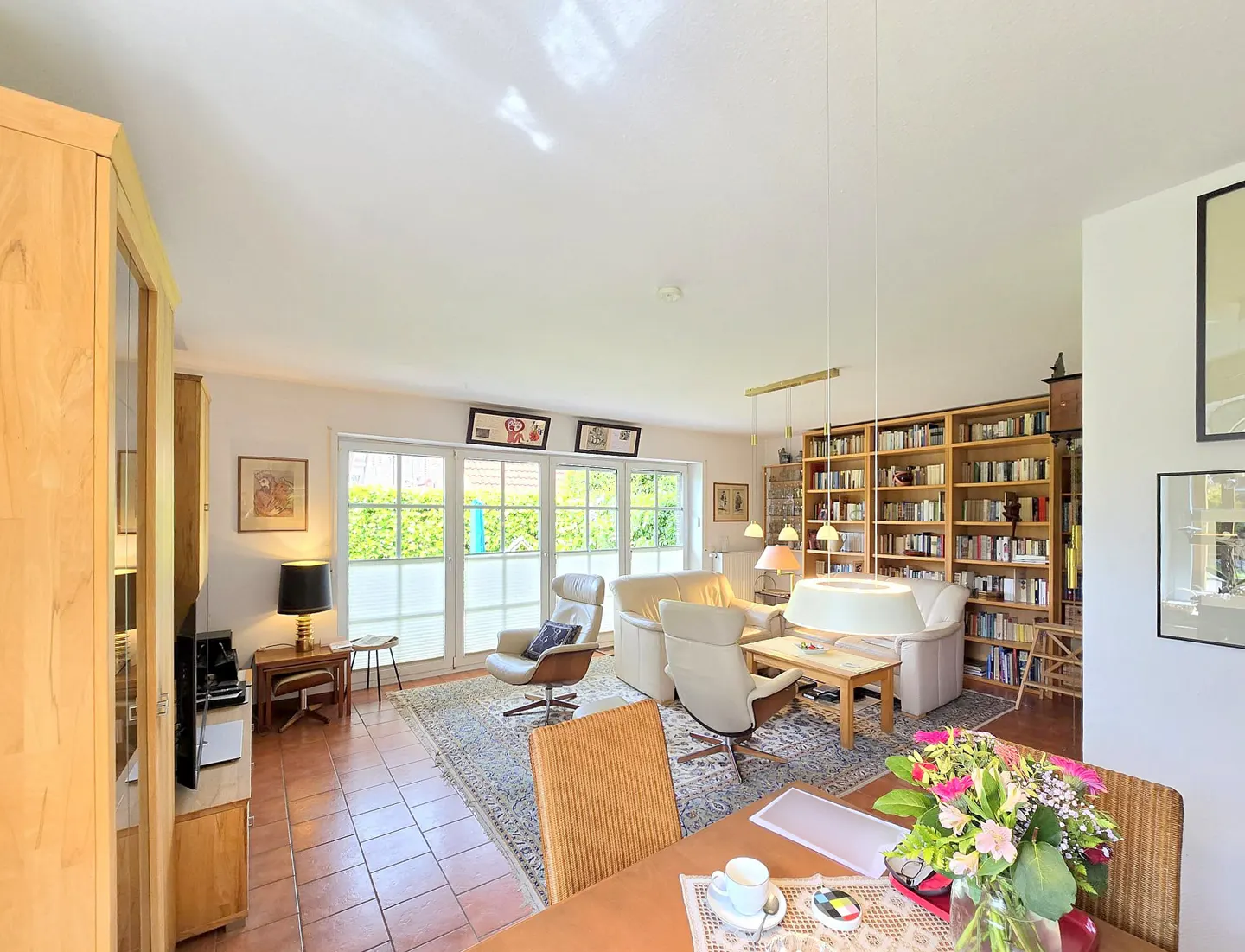 Bright living room with terracotta tile floor, white walls, and a large bookcase. A tan sofa and chairs sit on a patterned rug. French doors lead to a green yard.