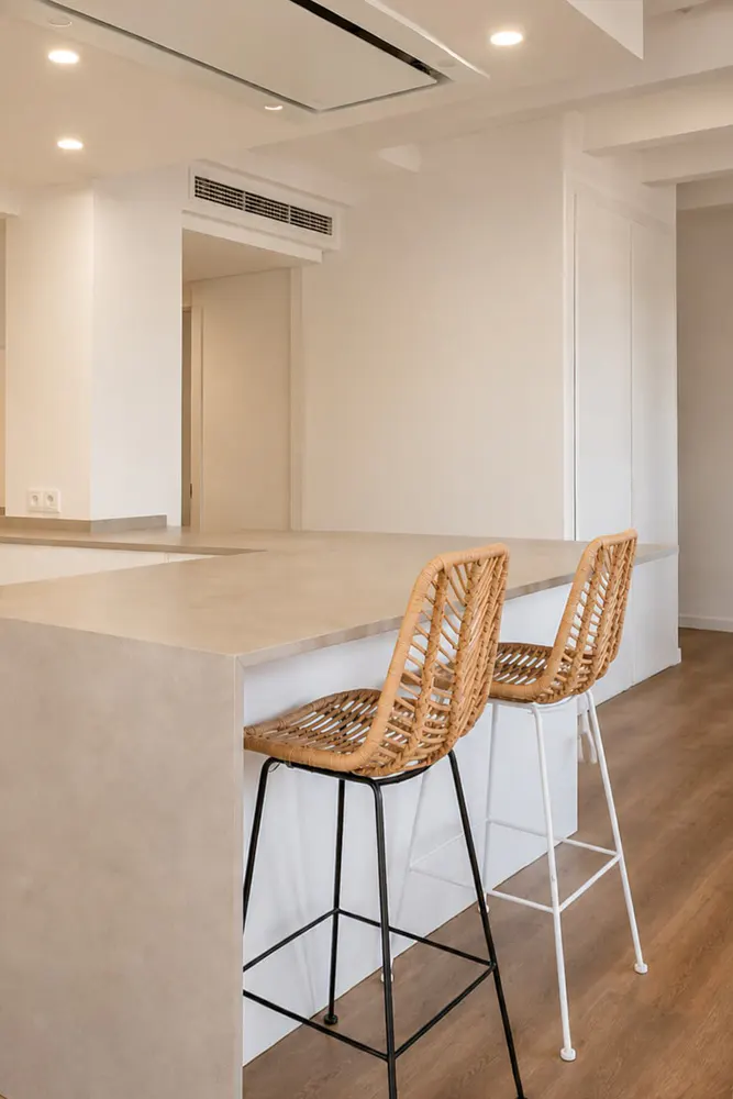 A bright, modern kitchen with two wicker bar stools at a light-colored island. White walls and wood floors create a clean, minimalist aesthetic.
