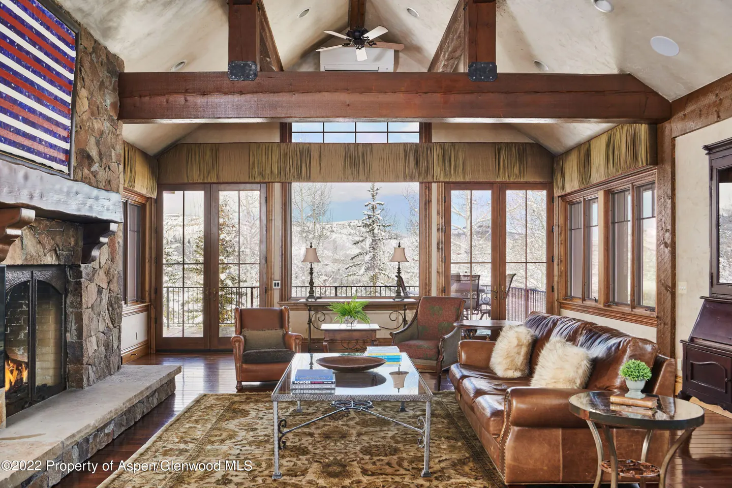 Living room with stone fireplace, leather furniture, and large windows overlooking a snowy landscape. Wooden beams accent the vaulted ceiling.