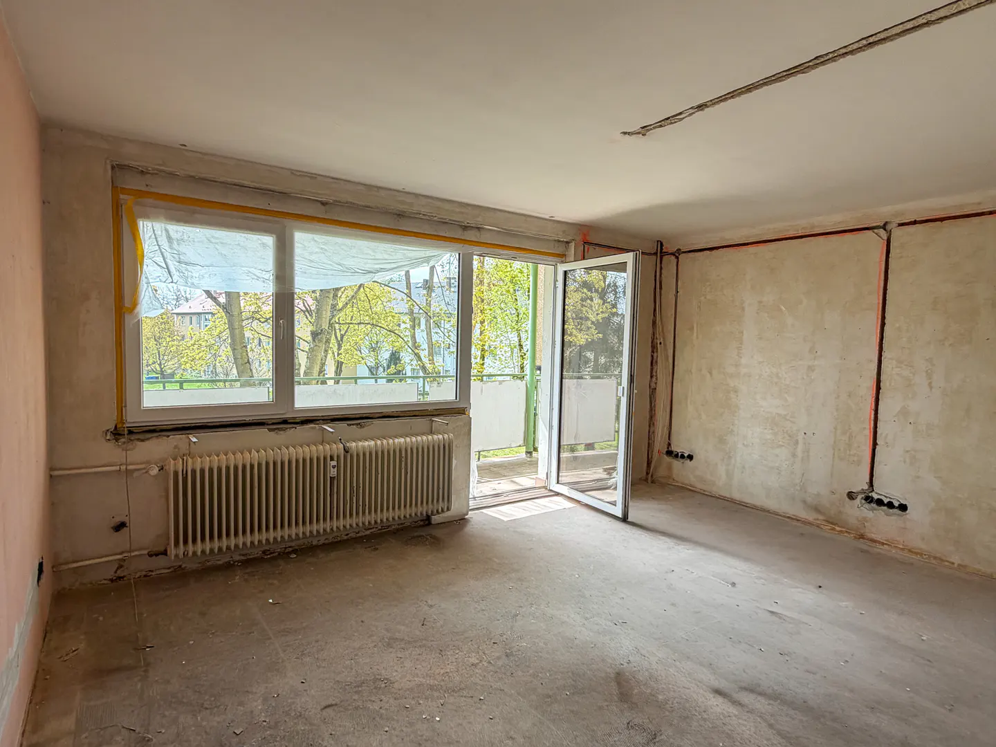 Unfinished room with open balcony door. Radiator under window with trees visible. Walls are bare with exposed wiring.