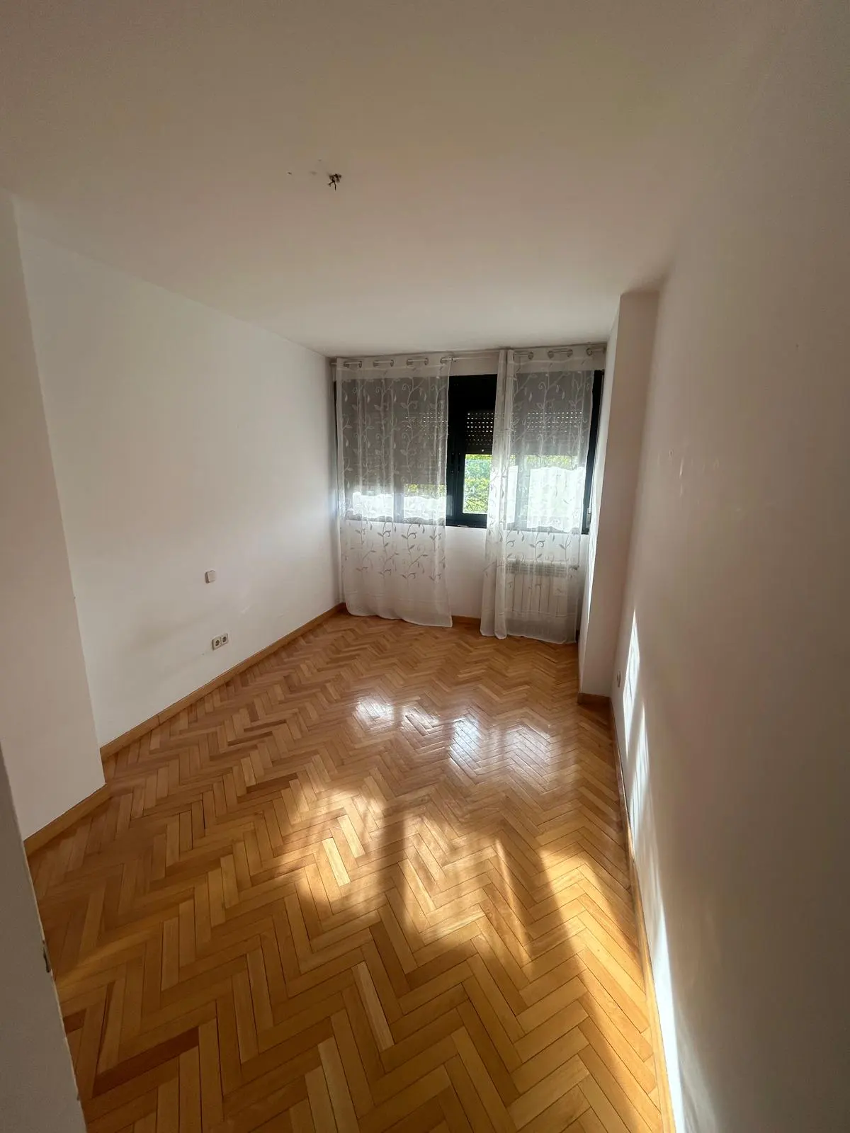 Bright, empty room with herringbone wood floors, white walls, and windows with sheer curtains.