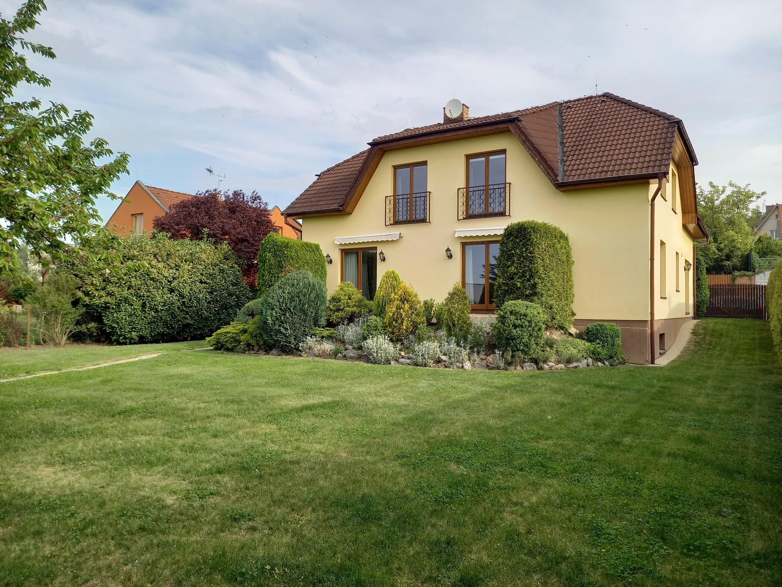 A two-story yellow house with a brown roof, balconies, and manicured green lawn and bushes.