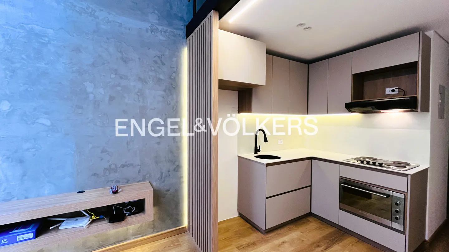 Modern kitchen with light beige cabinets, wood accents, and a concrete-textured wall with a floating shelf.