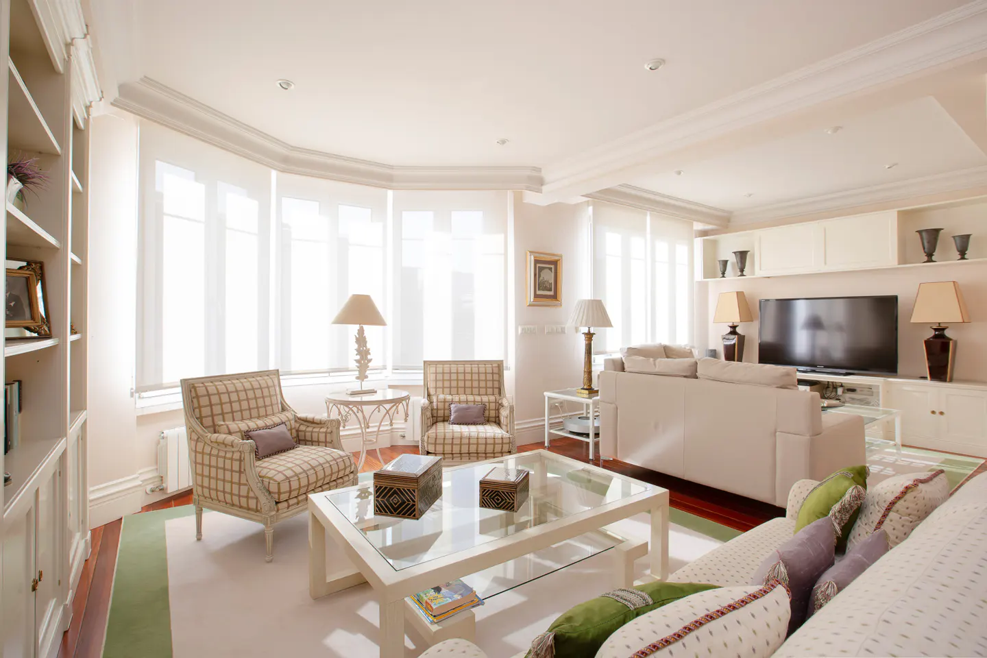Bright living room with white walls, large windows, and a glass coffee table. Two plaid armchairs face a white sofa. A TV sits on a built-in shelf.