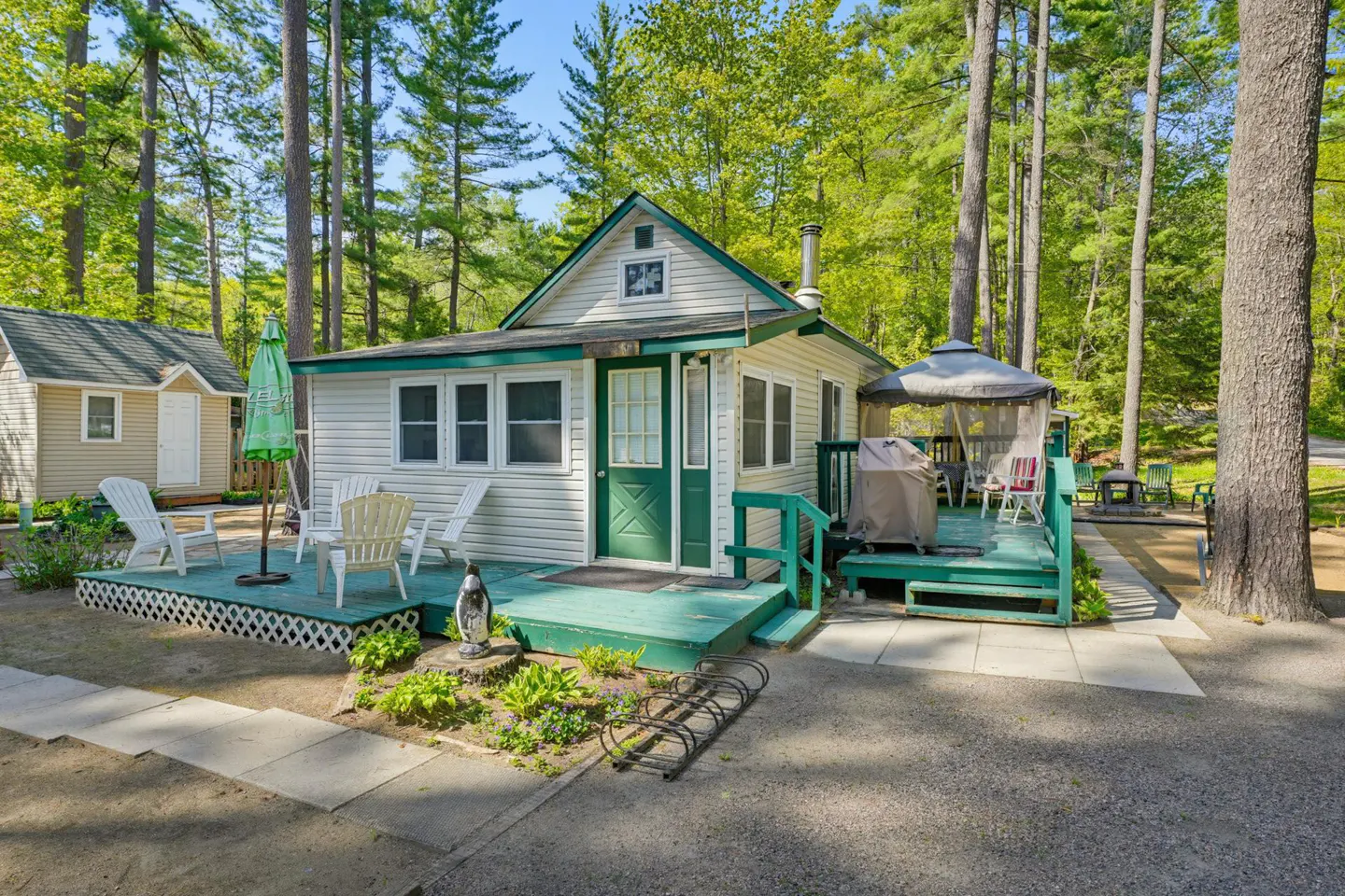 White cottage with green trim and a green deck, featuring outdoor seating, a gazebo, and surrounding forest trees.