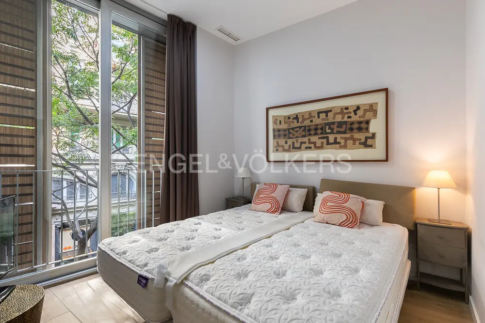 Bedroom with two twin beds, white walls, and a large window overlooking a tree-lined street. Artwork hangs above the beds.