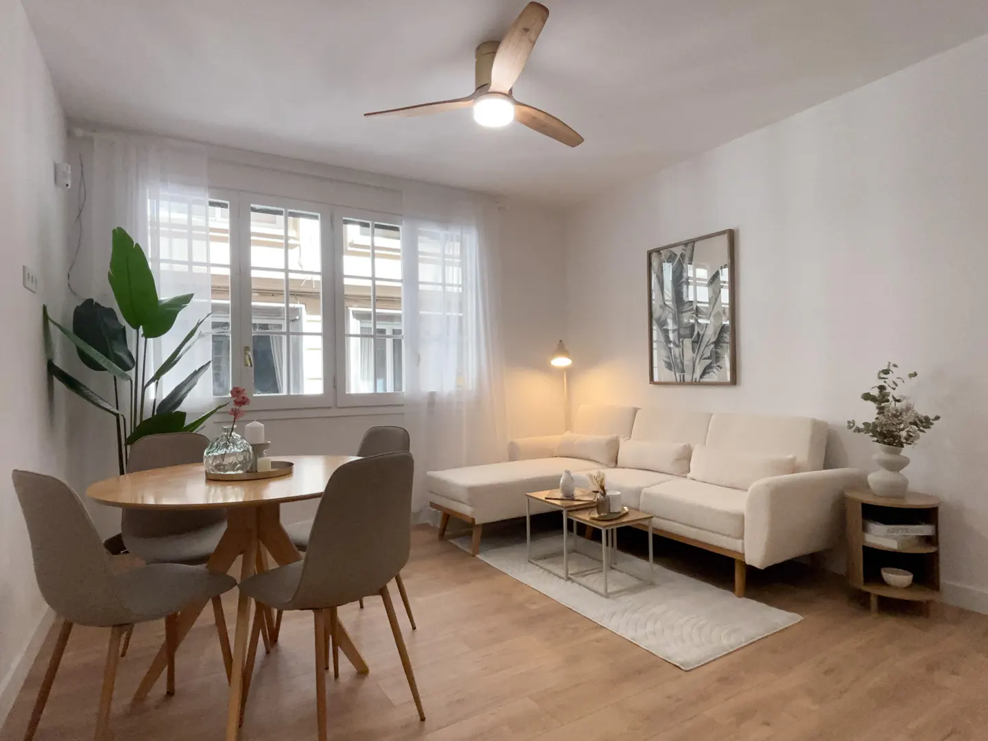 Bright living room with wood floors, a round table with chairs, and a white sectional sofa. A ceiling fan and large windows add to the airy feel.