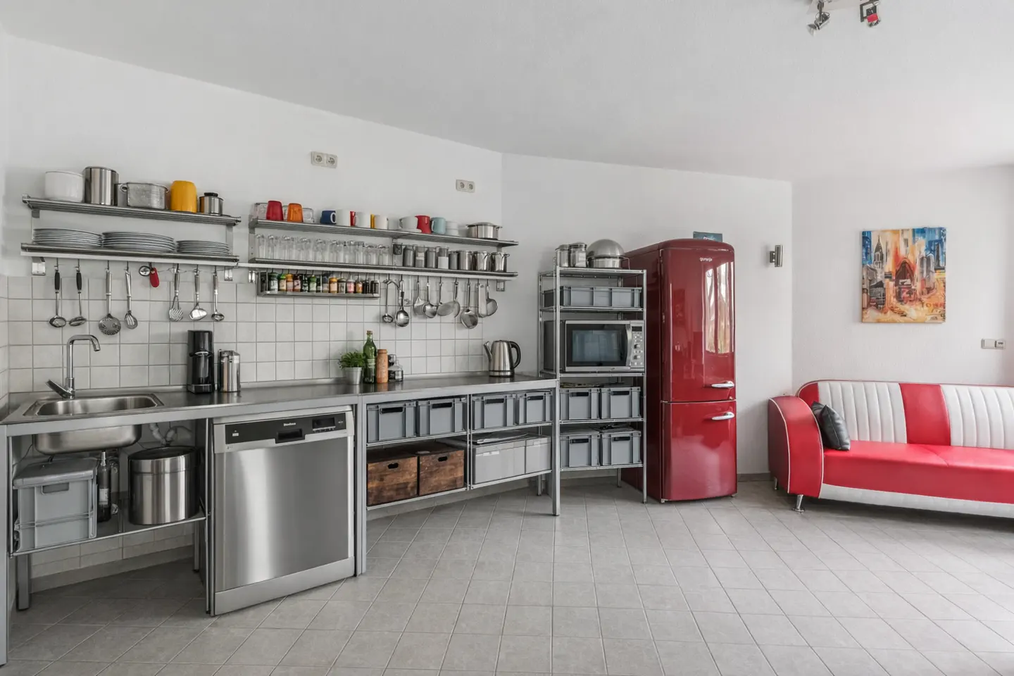 Bright kitchen with stainless steel counters, shelves, and appliances. A red retro fridge and red and white sofa add a pop of color.