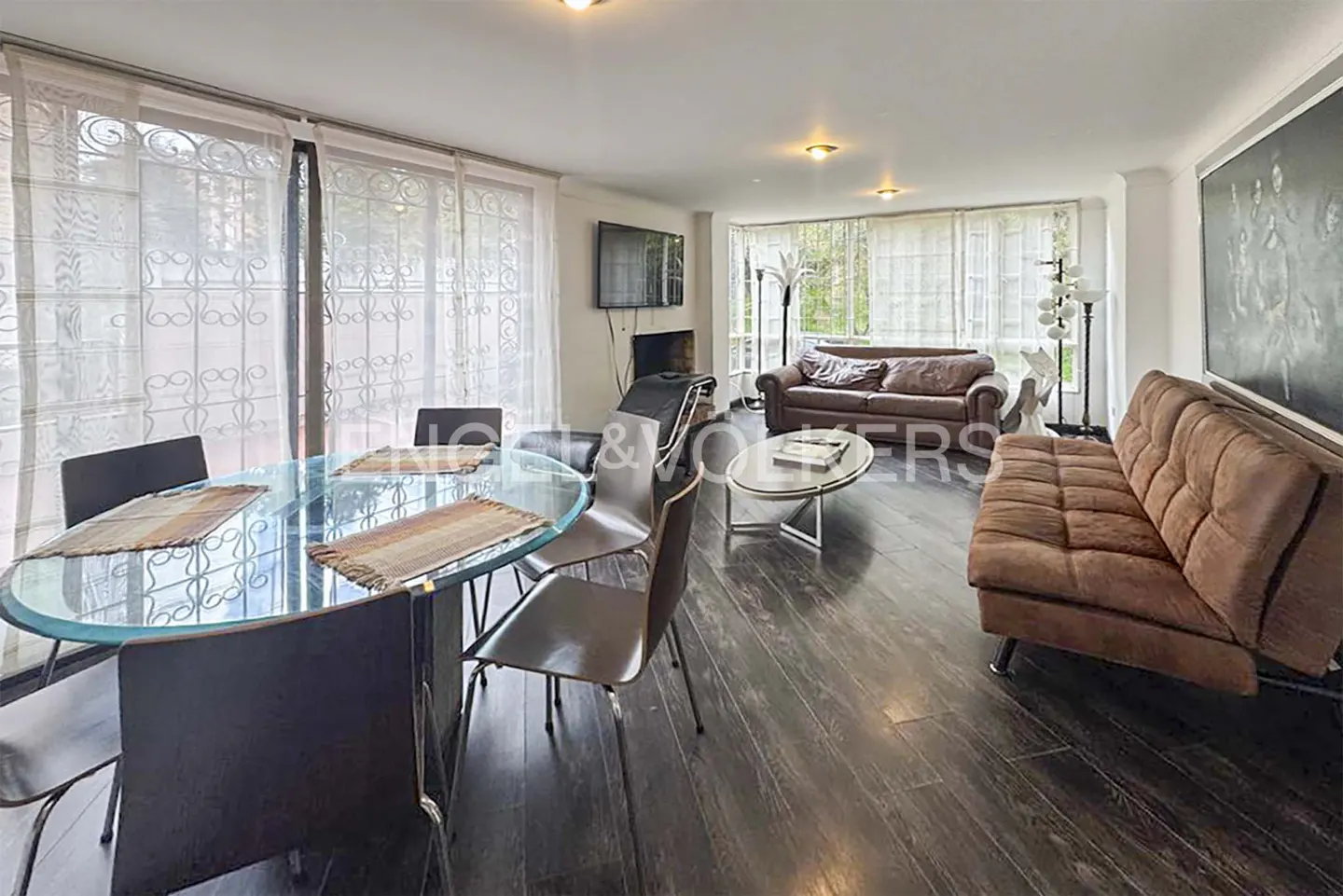 Open-concept living and dining area with a glass table, brown sofas, and dark wood floors. Natural light filters through sheer curtains.