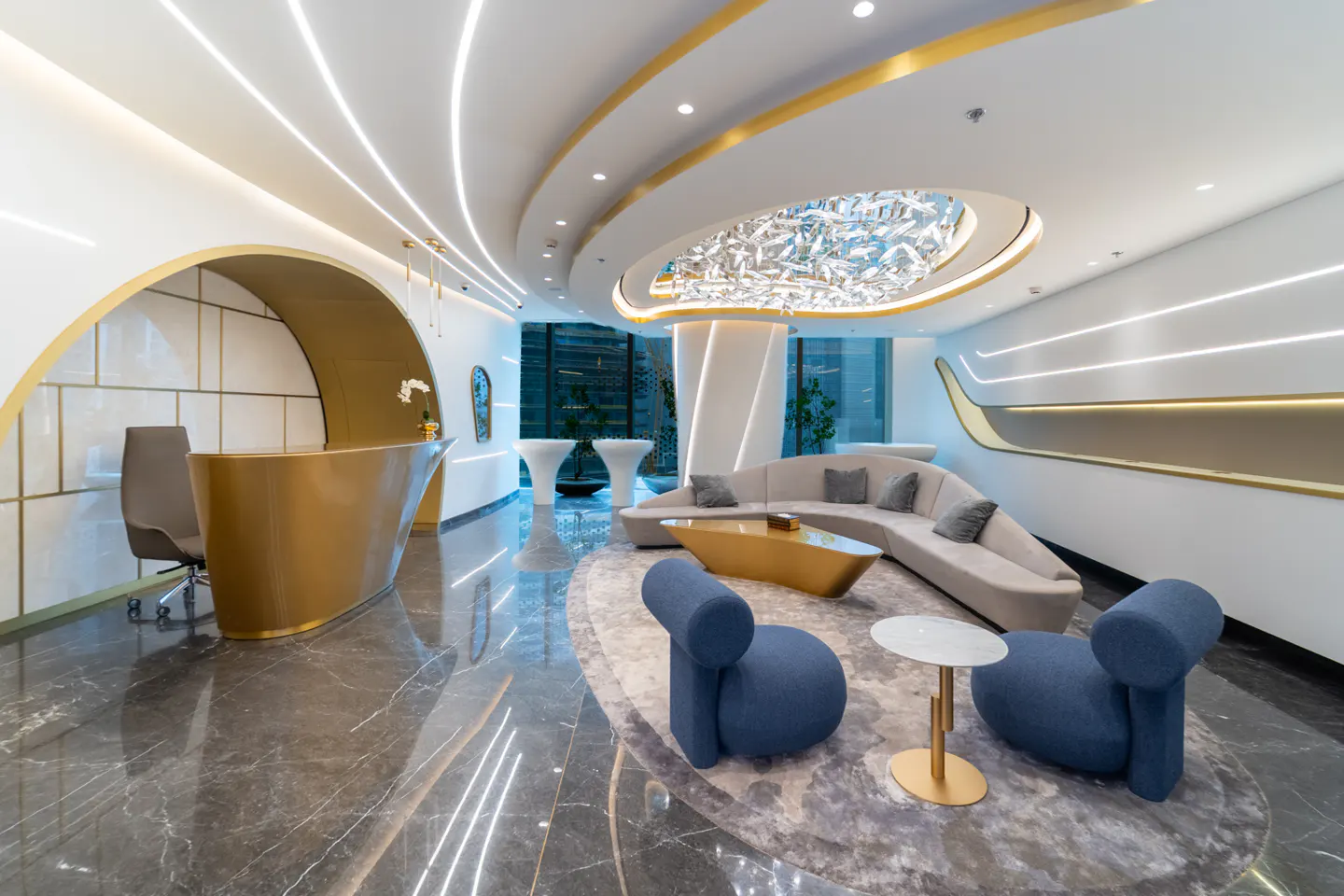 Modern lobby with gold reception desk, blue chairs, and curved sofa. A decorative light fixture hangs from the ceiling.