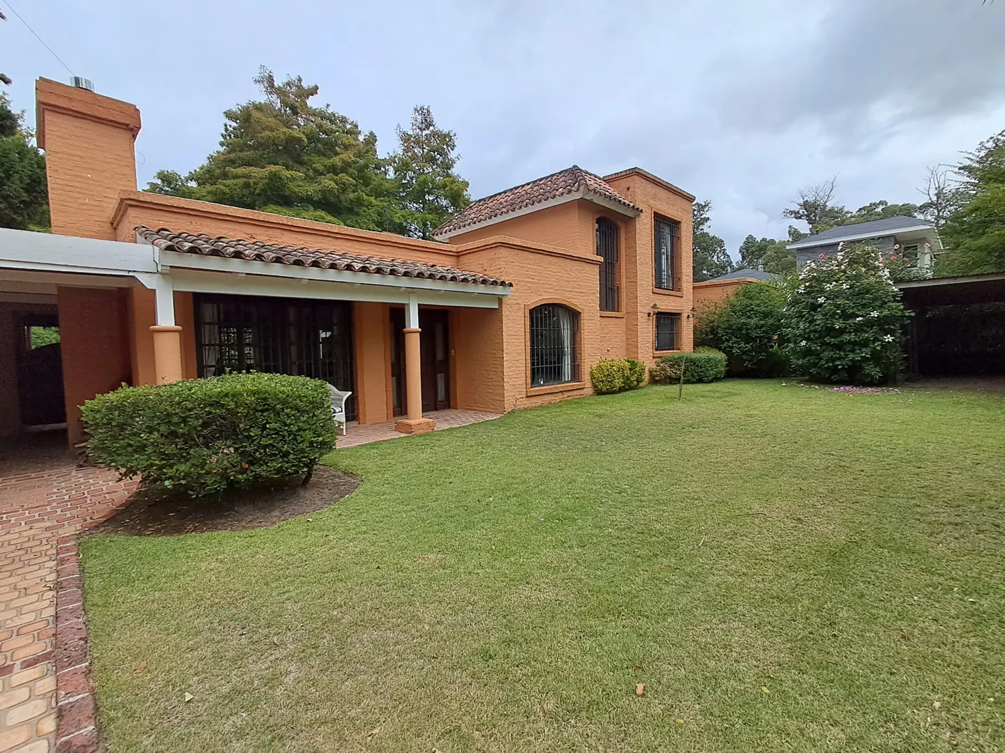 Two-story orange brick house with a tiled roof, covered patio, and a large green lawn under a cloudy sky.