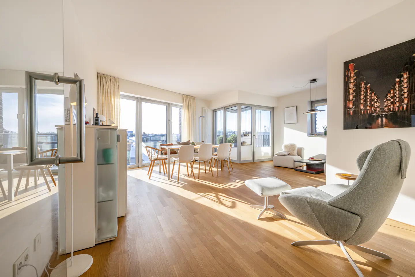 Bright, open-plan living room with light wood floors, a grey armchair, dining area, and large windows with natural light.