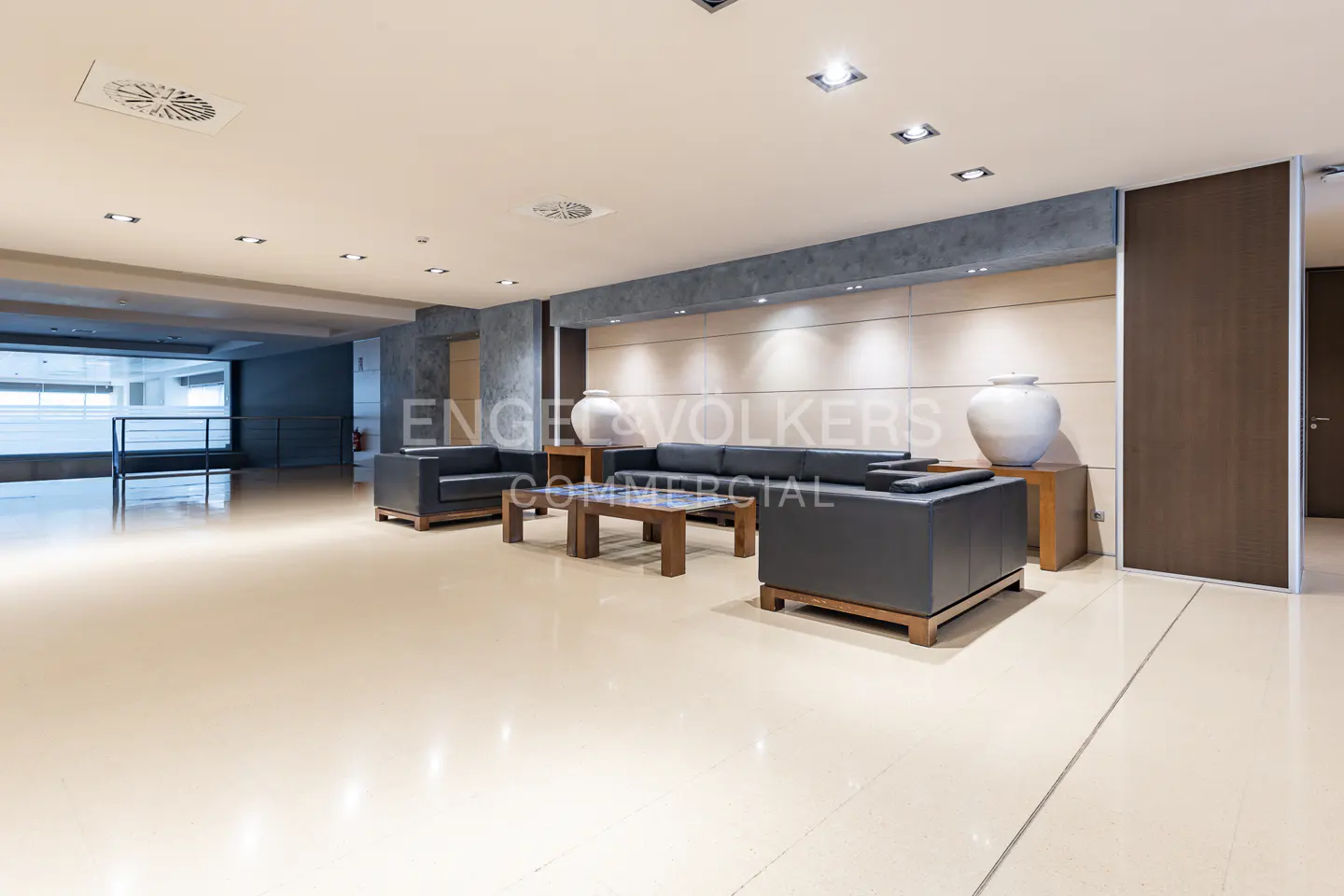 A modern lobby area with gray leather sofas, a wooden coffee table, and large white vases on wooden stands. The floor is a light beige color.