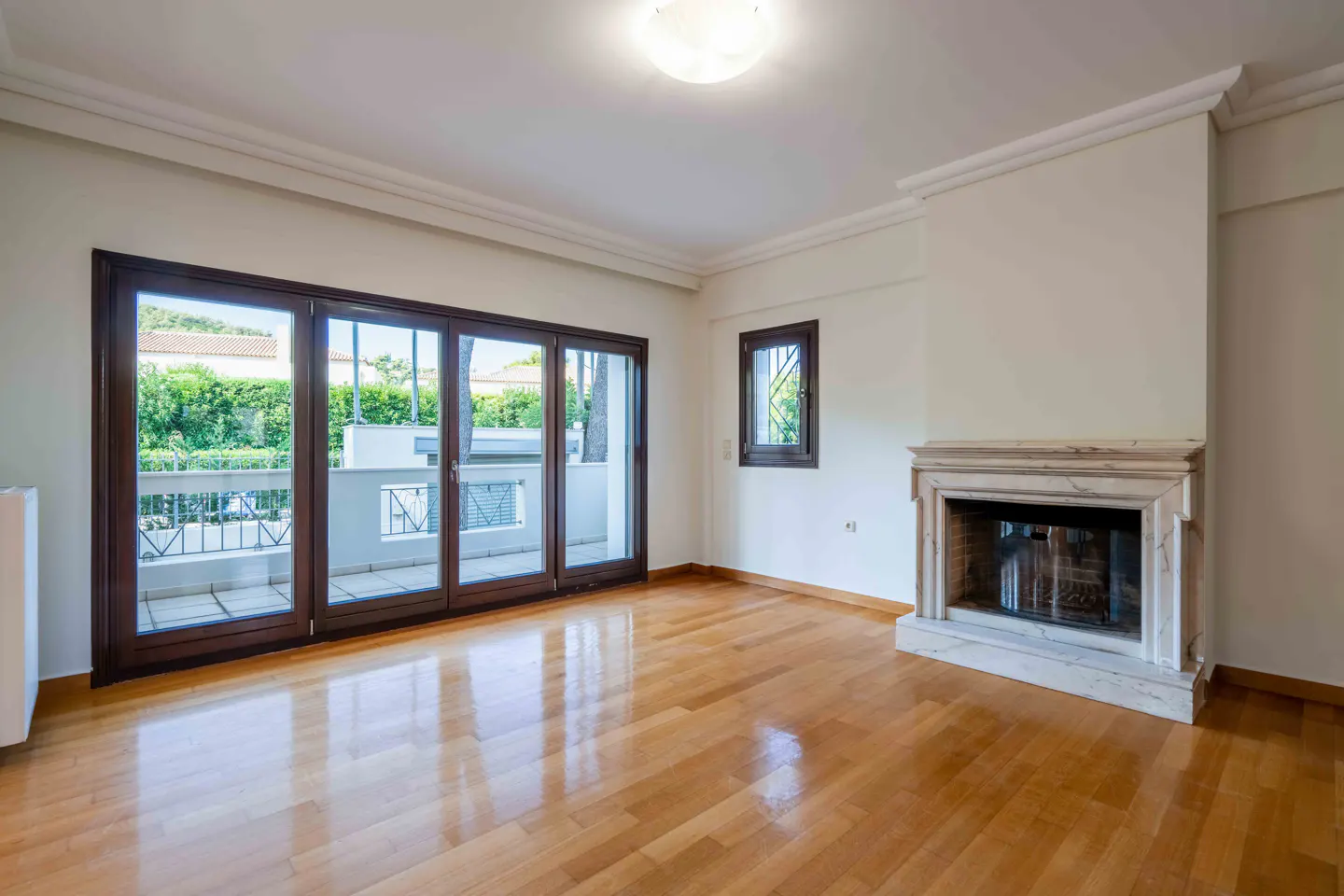 Bright, empty living room with hardwood floors, white walls, and a marble fireplace. Large, dark-framed glass doors lead to a balcony.
