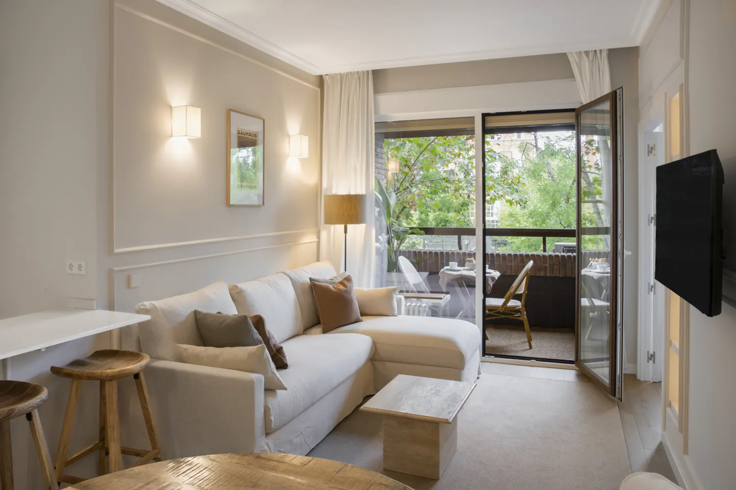 Living room with white sofa, wood stools, and balcony with table and chairs visible through open doors.