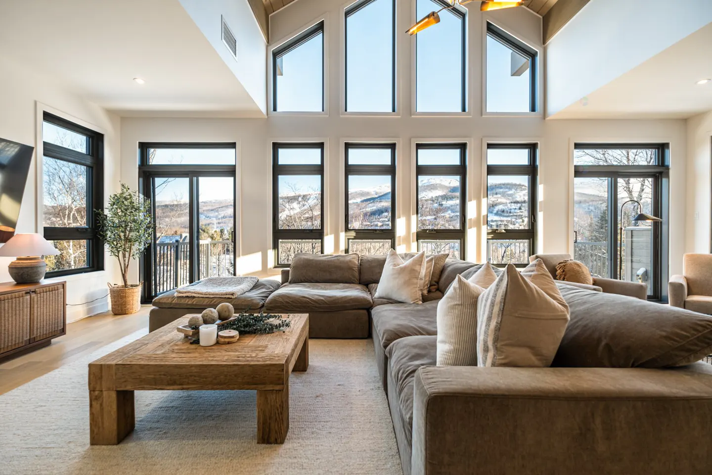 Bright living room with large windows showing a snowy mountain view. A brown sectional sofa and wooden coffee table sit on a light rug.