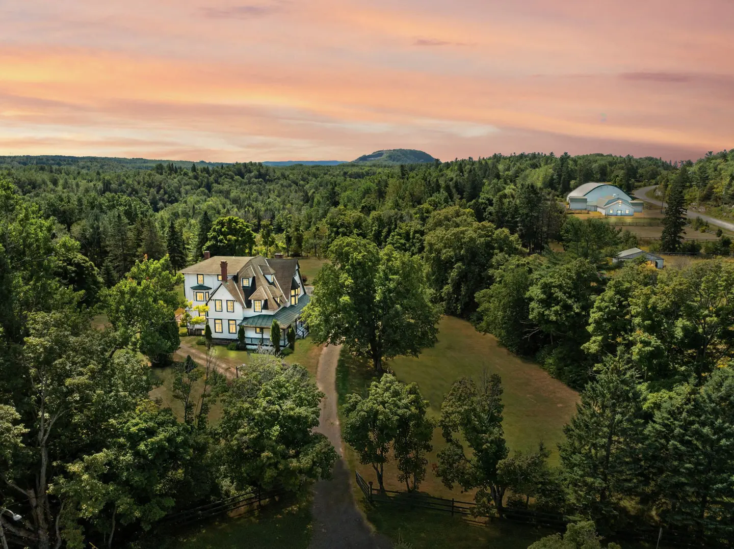 Aerial view of a large white house surrounded by green trees under a pink and orange sky.