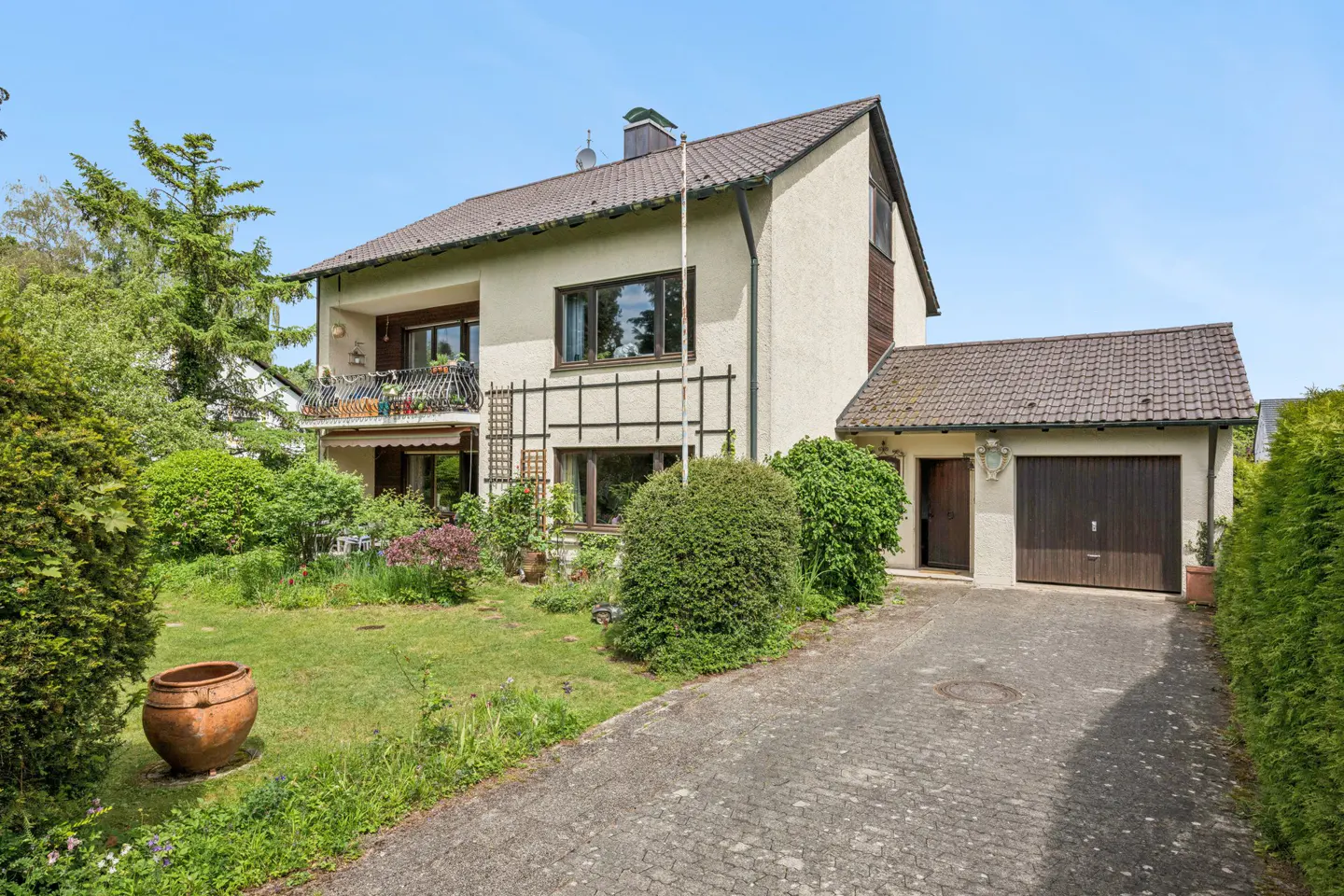 Two-story beige house with a brown tiled roof, attached garage, paved driveway, and a lush green front garden.