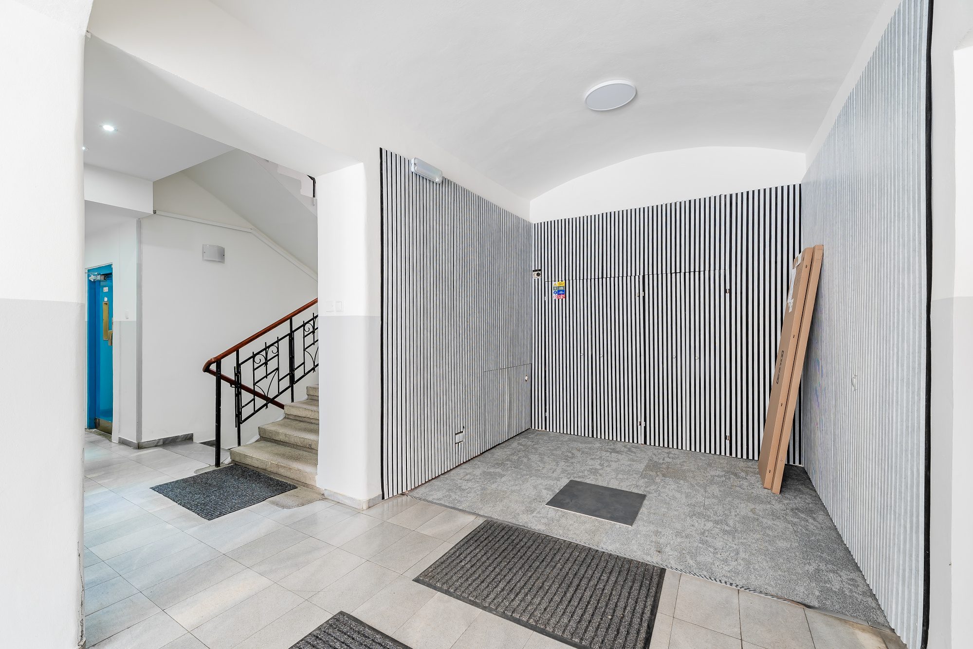 Modern apartment building lobby with black and white striped walls, stone flooring, and a staircase with a wooden railing.