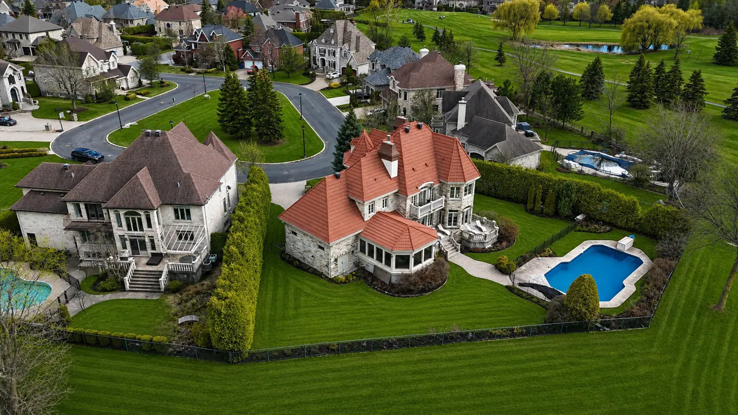 Aerial view of luxury homes with manicured lawns, swimming pools, and a golf course in the background.