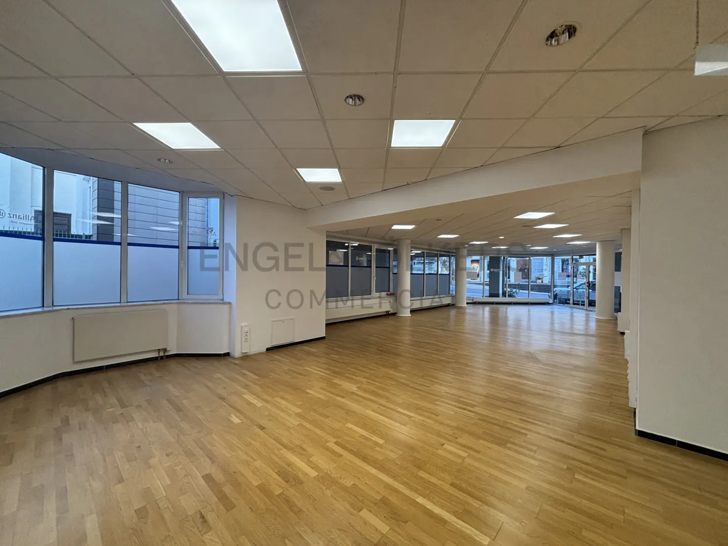 Empty commercial space with light wood floors, white walls, and large windows. Recessed lighting in the white ceiling.