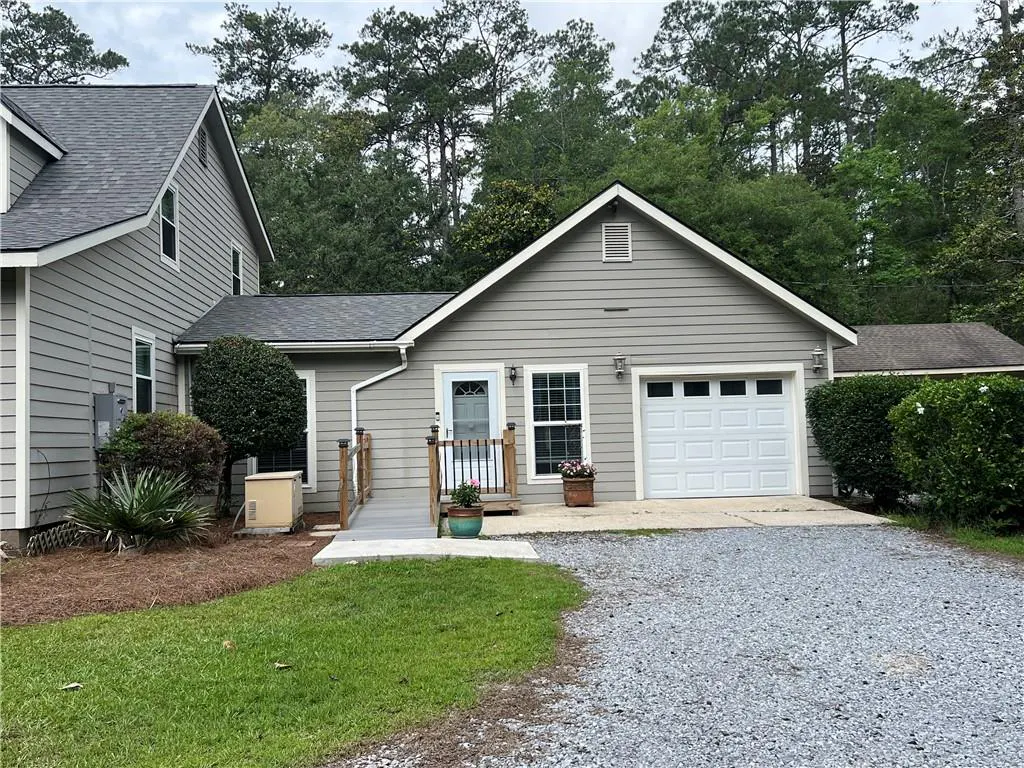 Exterior view of a gray, two-story house with a white garage door and a ramp leading to the front door. The yard has grass and gravel.