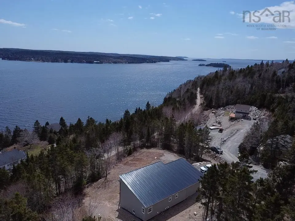 Aerial view of a gray house with a metal roof, surrounded by trees, overlooking a blue ocean on a sunny day.