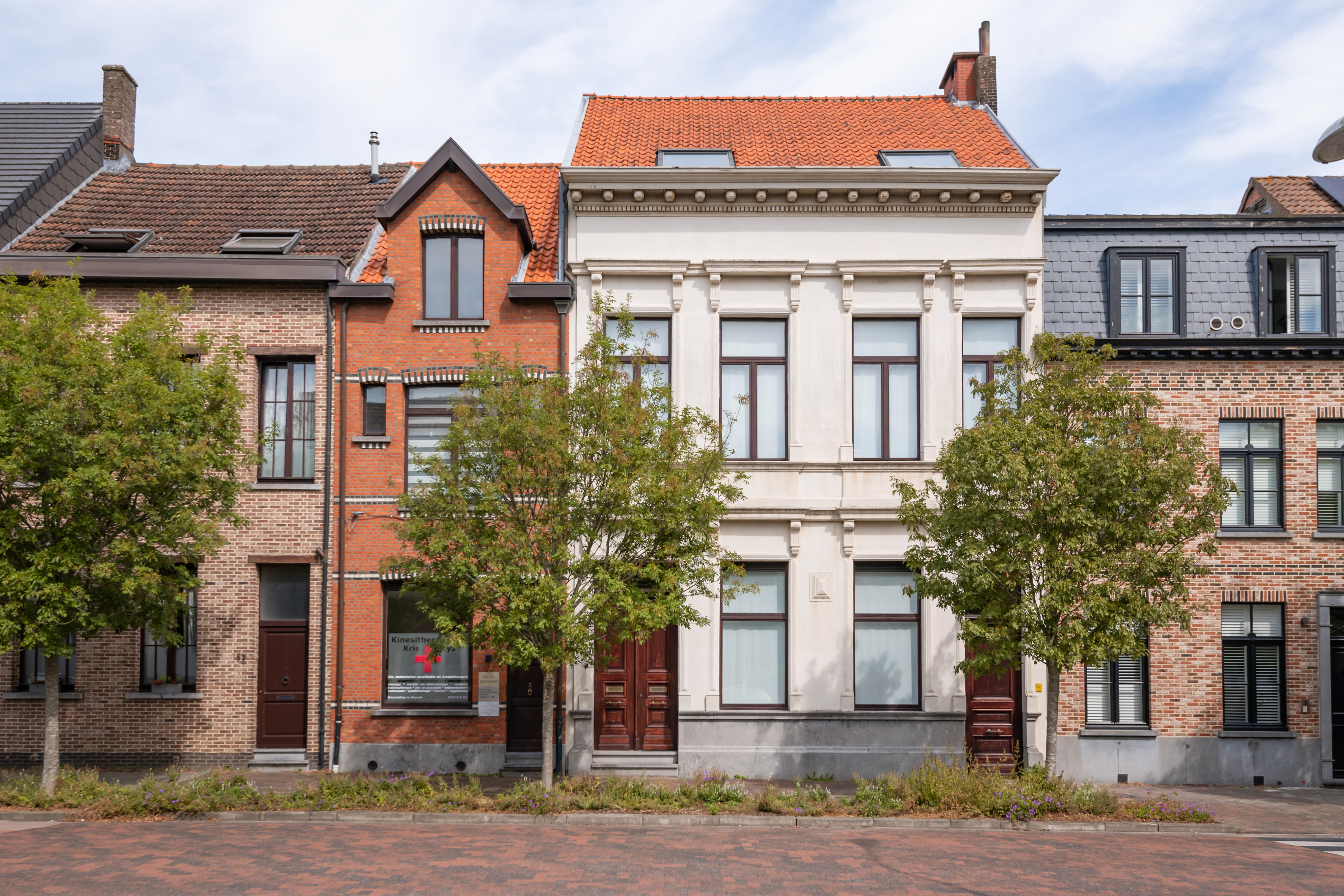 A row of historic European townhouses with brick and cream facades, tall windows, and green trees lining the sidewalk. [AI-generated text]
