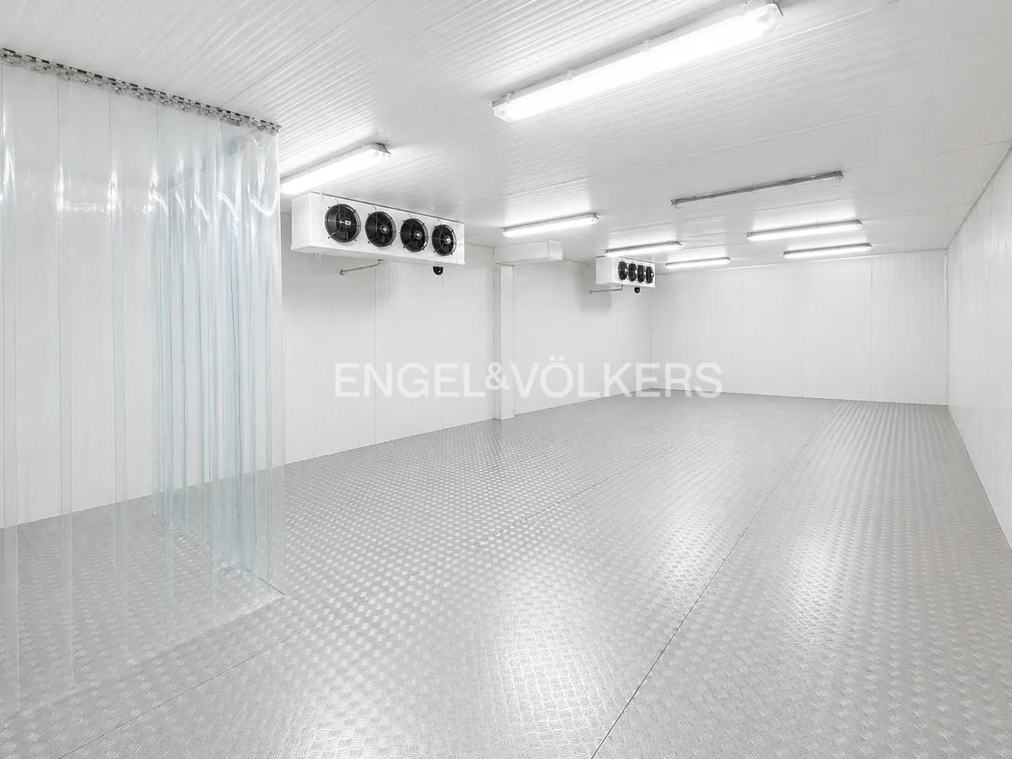 Bright, empty commercial freezer with white walls, diamond-patterned floor, plastic strip door, and Engel & Völkers logo.