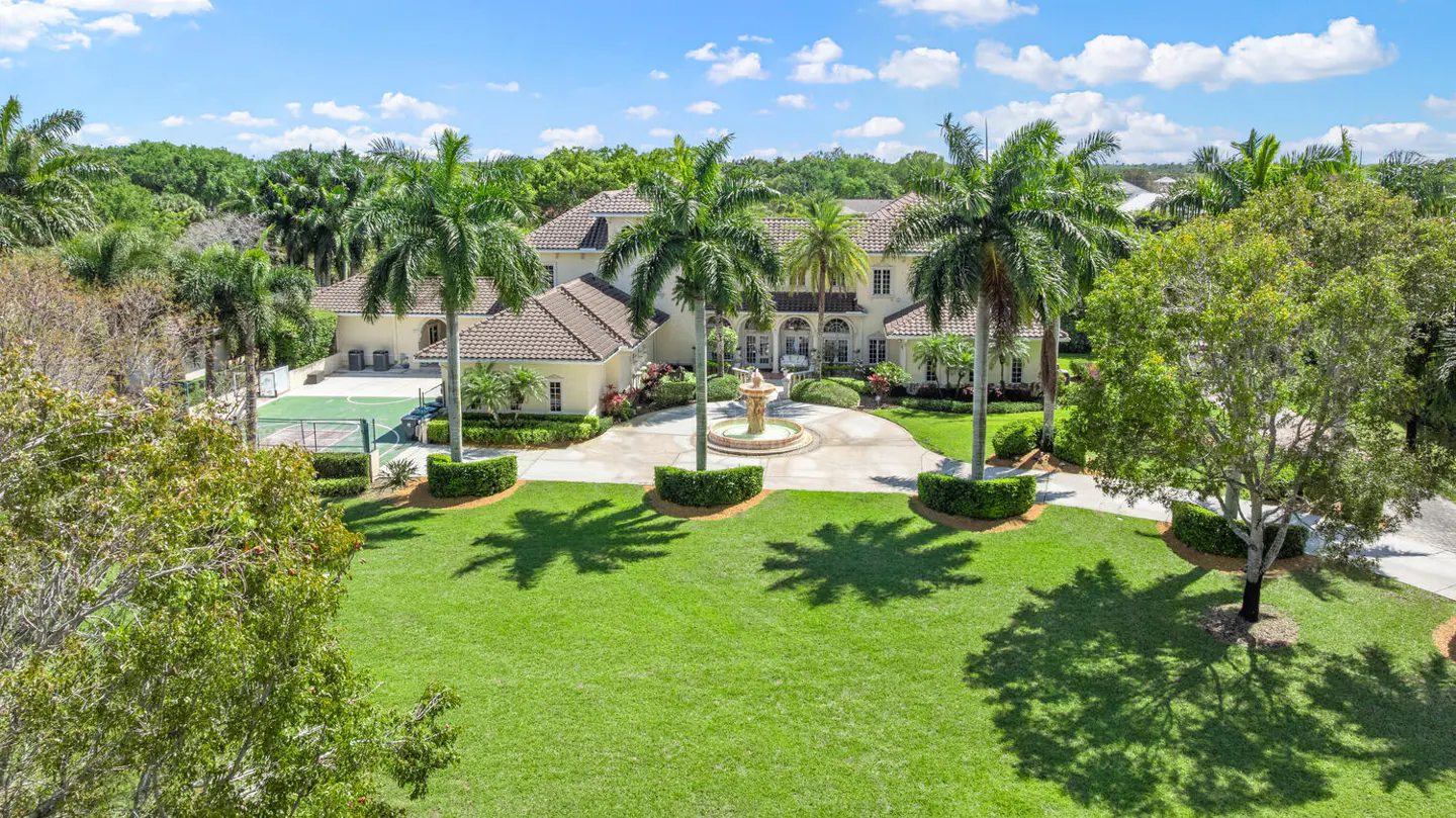 Beige mansion with a brown tile roof, palm trees, a fountain, and a green lawn on a sunny day.