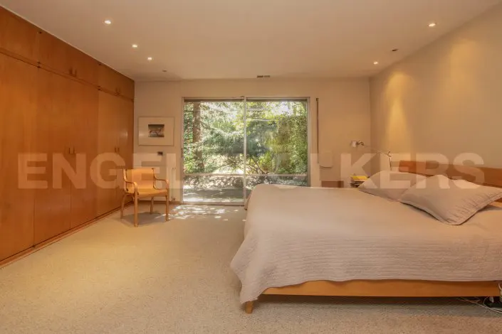 Bedroom with a large bed, wood closet, chair, and sliding glass doors to a green outdoor space.