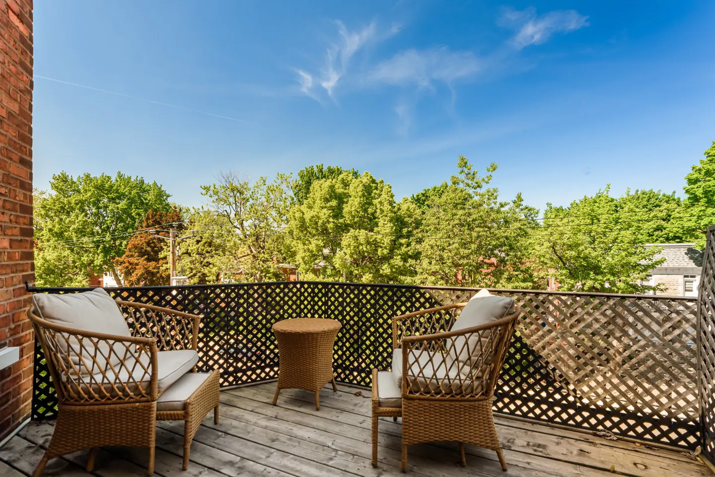 A balcony with two wicker chairs, a small table, and a lattice railing overlooking green trees under a blue sky.
