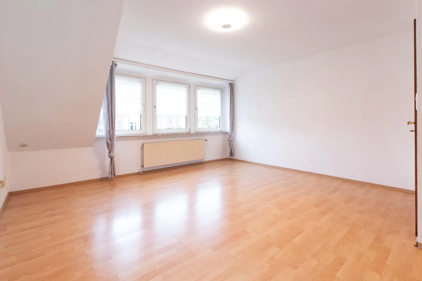 Bright, empty room with wood floors, white walls, and three windows with gray curtains. A radiator is under the windows.
