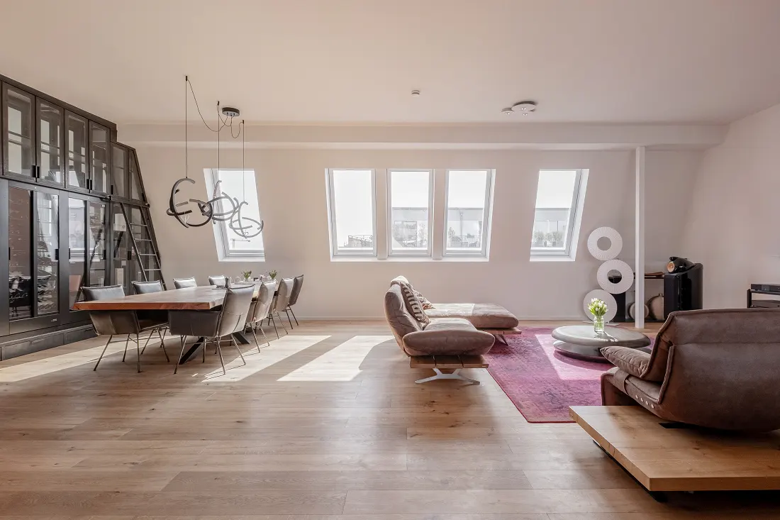 Bright, modern living space with wood floors, a long dining table, and a brown leather sofa on a pink rug. Skylight windows let in natural light.