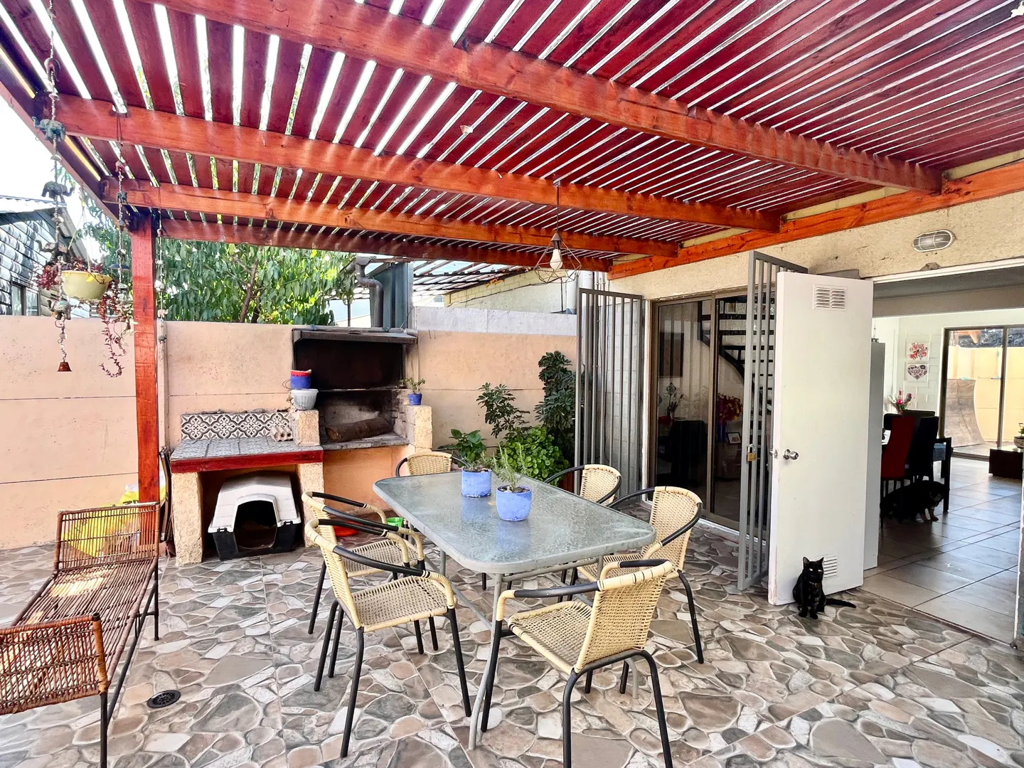 Outdoor patio with a wooden pergola, stone floor, grill, table with chairs, and a black cat sitting near the open door.