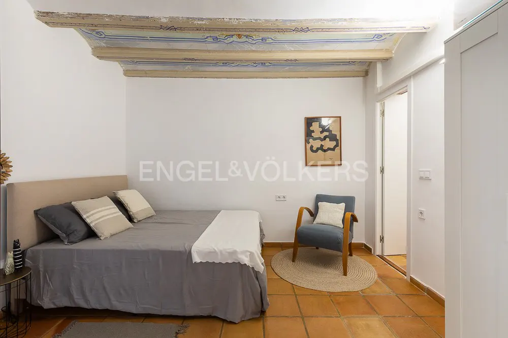 Bedroom with a gray bed, blue chair on a round rug, and terracotta tile floor. The ceiling has decorative beams. A framed print hangs on the white wall.